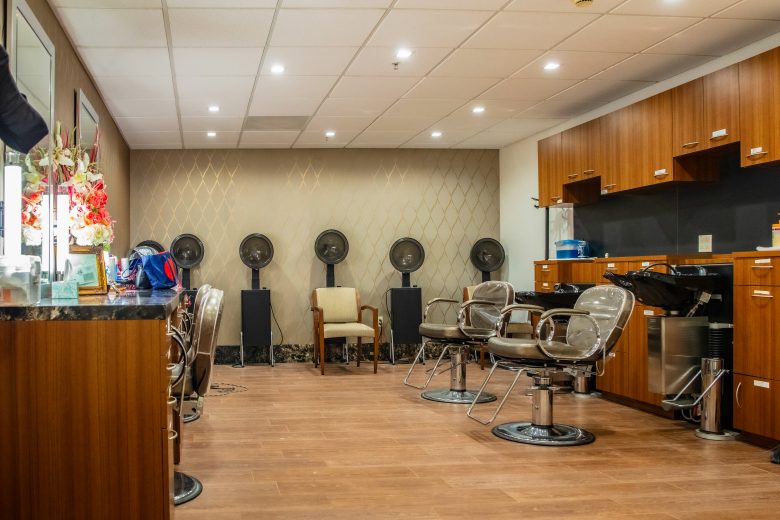 A modern hair salon interior with brown wooden floors, salon chairs, wash sinks, and hairdryers in the back row creates a chic lifestyle vibe. Cabinets line the right wall, while decorative flowers brighten the counter to the left.