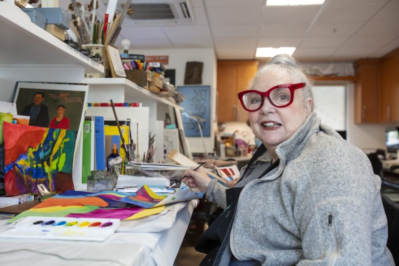 An older woman with gray hair and red glasses sits at a table painting with watercolors in a bright art studio, surrounded by colorful artwork and art supplies. She smiles at the camera.
