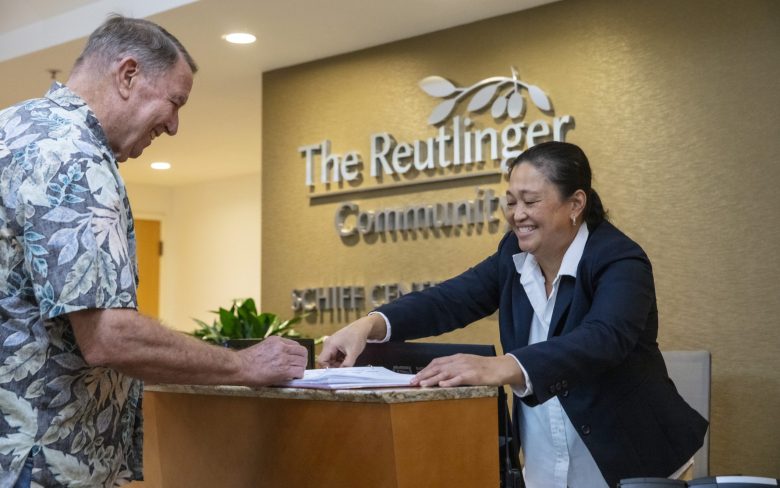 A man in a floral shirt signs papers at a reception desk while a smiling woman in a blazer assists him. The background displays a sign reading The Reutlinger Community.