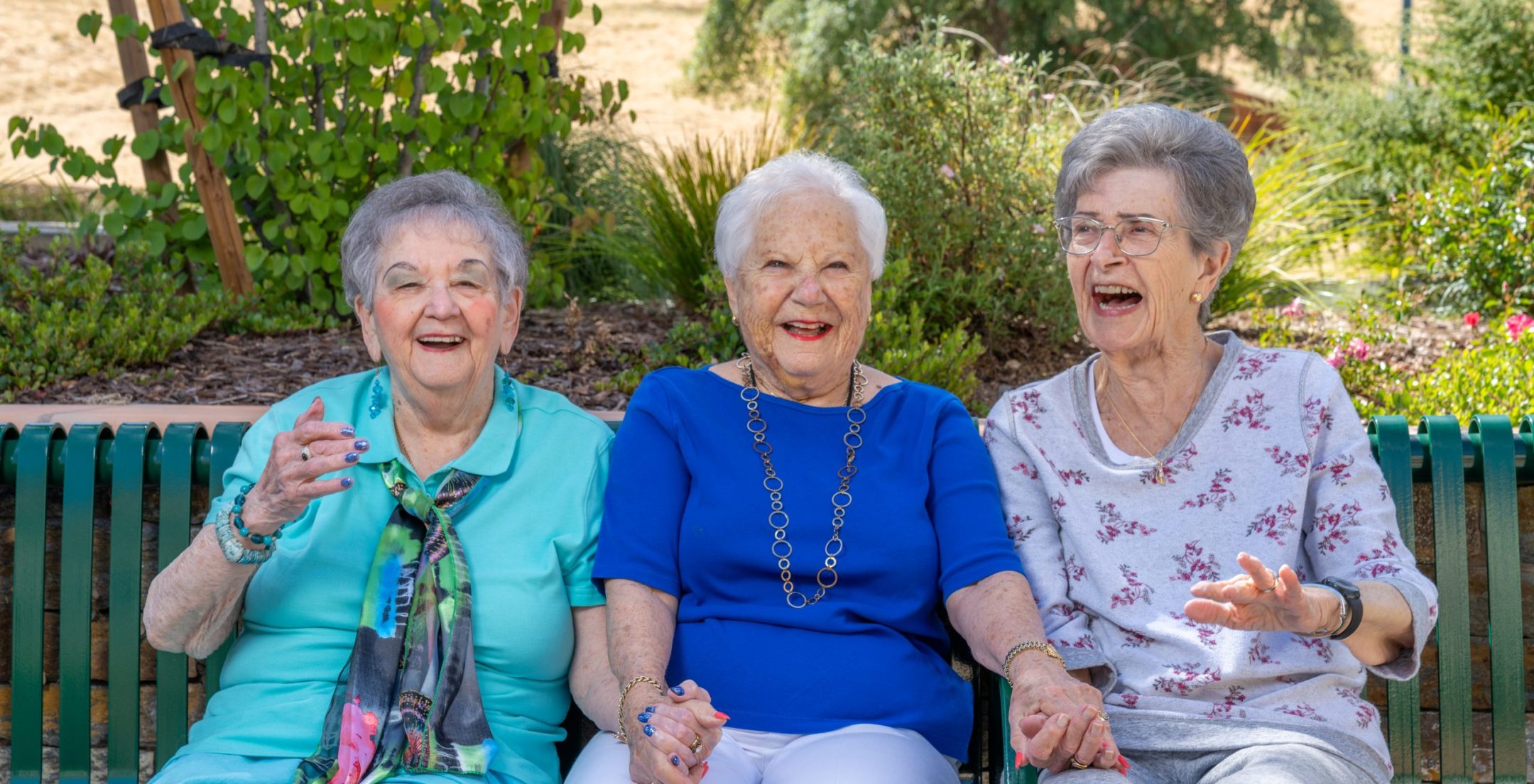 Three elderly women sit on a green bench outdoors, smiling and laughing together as they discuss living options. The background features greenery and trees, suggesting a peaceful park or garden setting.