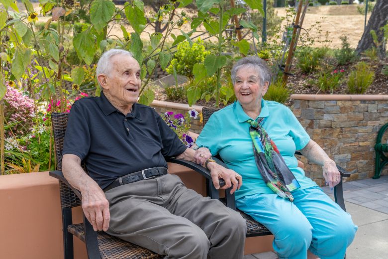 Two elderly people sit holding hands and smiling on patio chairs in a garden, surrounded by greenery and flowers, enjoying a sunny day outdoors while exploring different living options.