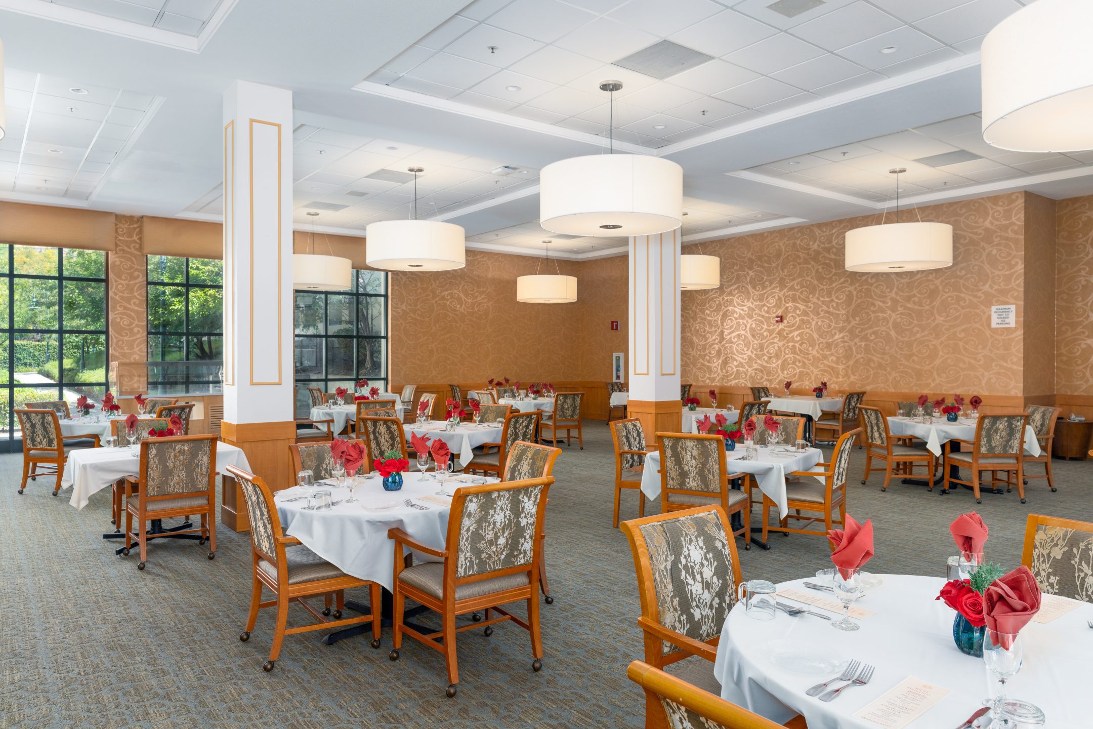 Elegant dining room with round tables set for a meal, each covered with white tablecloths and red napkins. Wooden chairs have patterned cushions. Large windows and modern pendant lights enhance the resident experience.