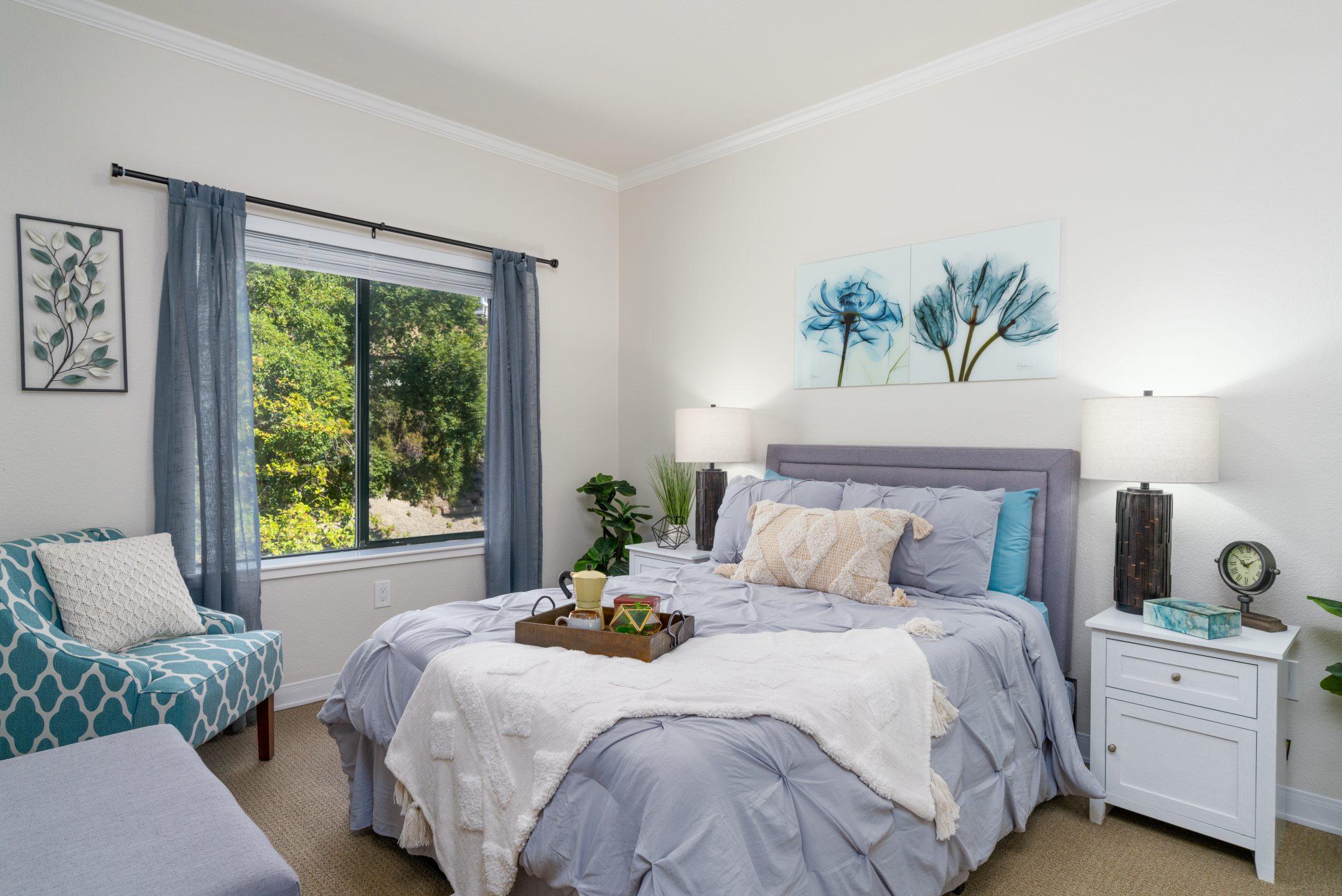 A cozy bedroom with a neatly made bed in soft gray bedding, two bedside tables with lamps, a window with blue curtains, and a patterned armchair—one of the comfortable Living Options that also features wall art and greenery visible outside.