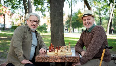 Two older men sit at a park bench, smiling at the camera with a chessboard between them. Trees and greenery are visible in the background, and one man holds a cane.