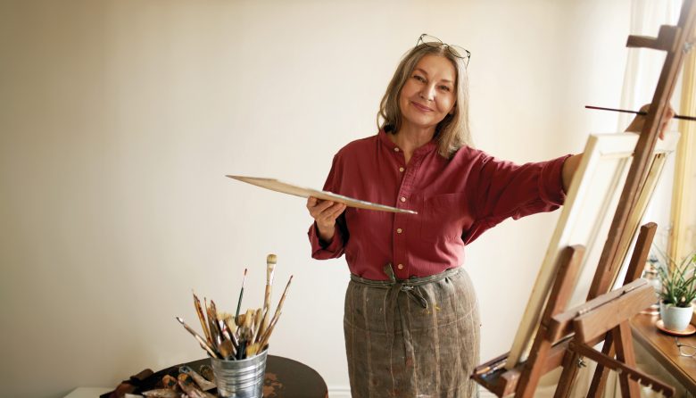 A woman in a red shirt and apron stands at an easel, smiling while holding a palette. Art supplies and brushes are on a table nearby in a bright room with light walls.