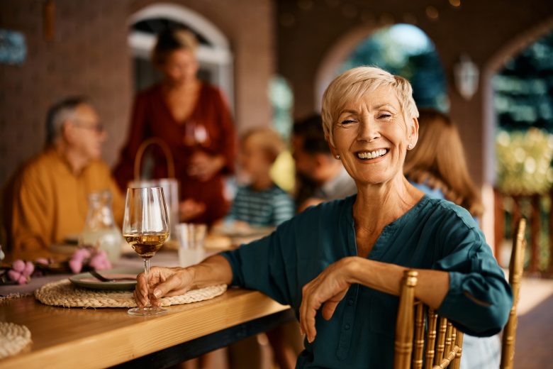 A smiling older woman with short blond hair sits at an outdoor table holding a glass of white wine, with a group of people gathered and talking in the background.