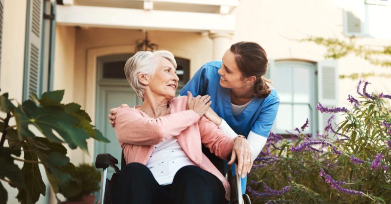 An elderly woman in a wheelchair smiles and holds hands with a caregiver in blue scrubs. They are outdoors near a house, surrounded by purple flowers, sharing a warm Skilled Nursing moment filled with care and compassion.
