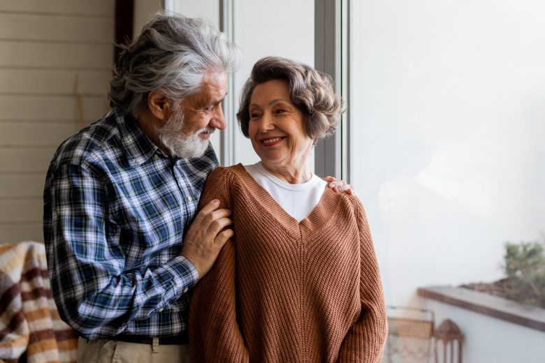 An elderly couple stands by a window, looking at each other and smiling warmly. The man gently rests his hand on the womans shoulder. They appear happy and comfortable together in a cozy indoor setting.