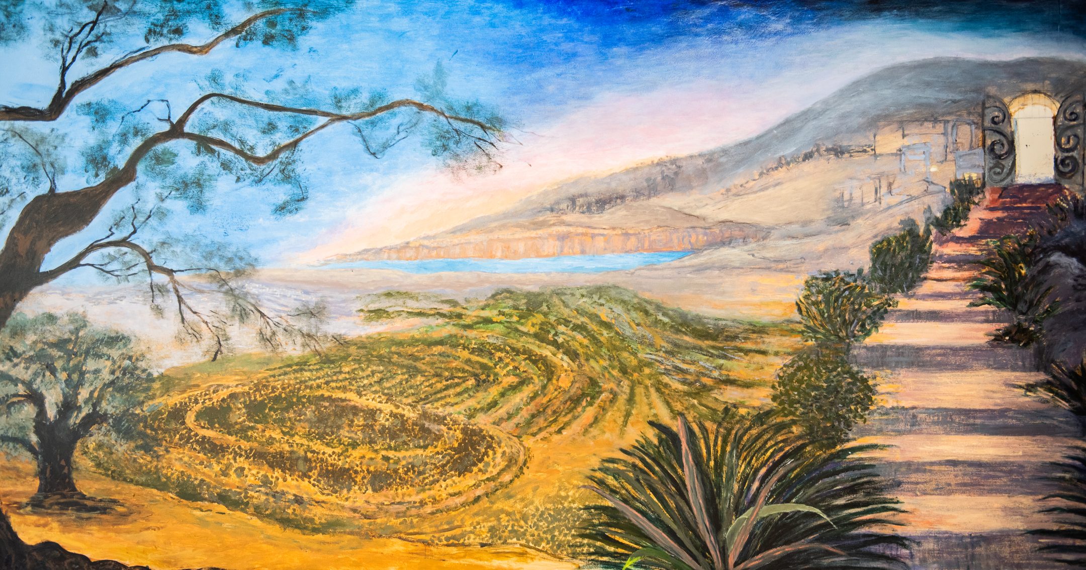 A painted landscape featuring rolling fields, a winding path lined with plants, a tree on the left, distant mountains, a body of water, and a gate leading to a building on a sunlit hillside under a blue sky.