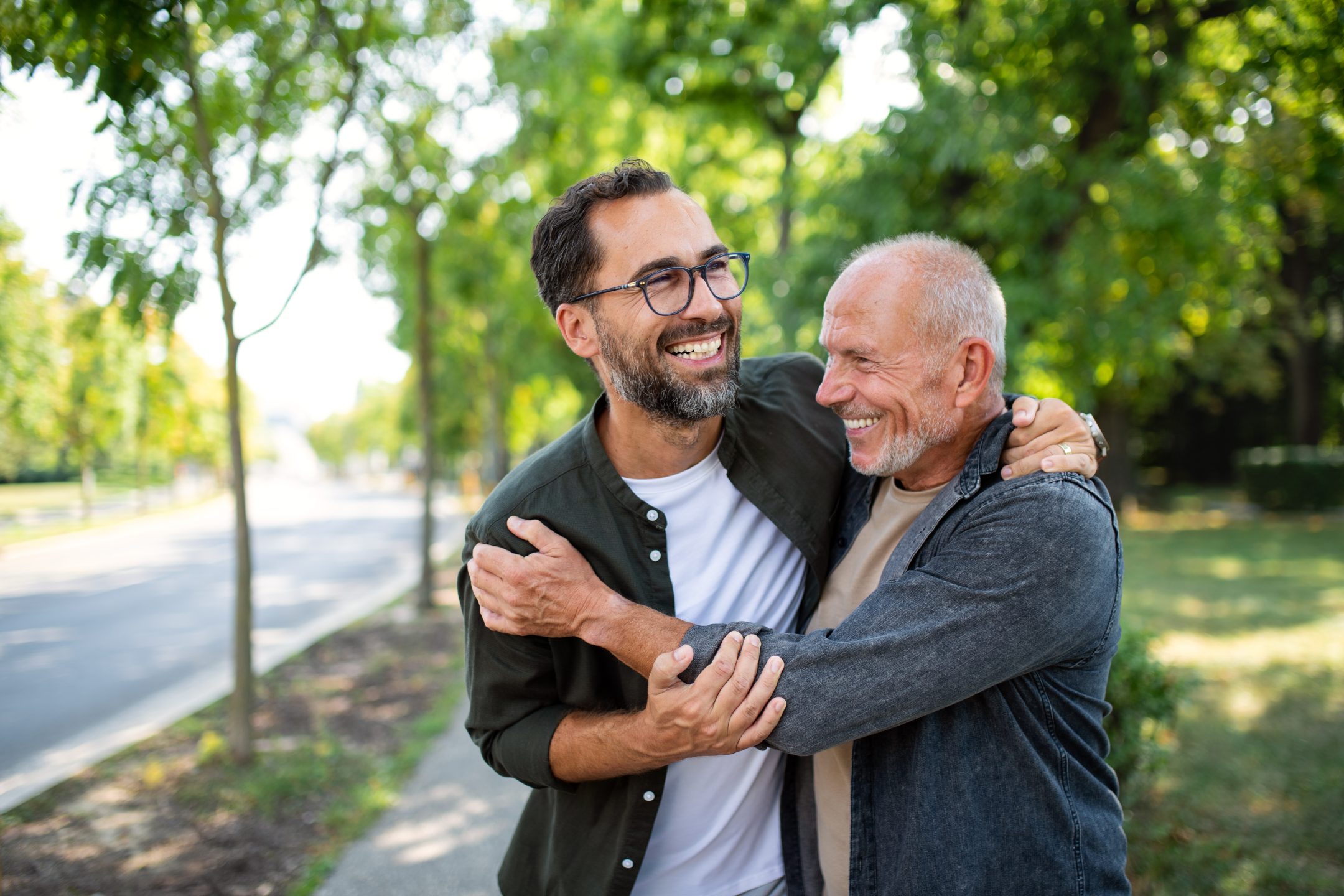 Two men standing on a tree-lined sidewalk, smiling and embracing each other in a joyful, affectionate moment on a sunny day.