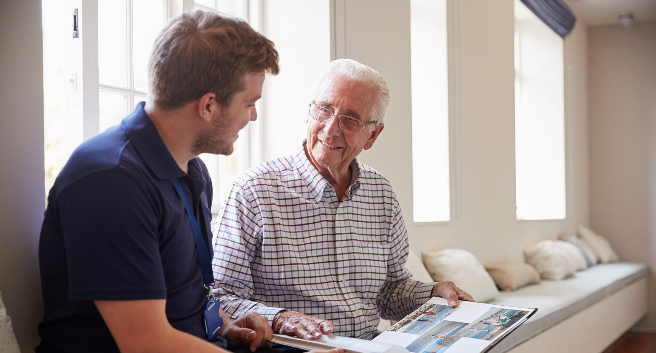 A young man and an older man sit by a bright window at The Reutlinger memory care Danville CA, looking at a photo album together and smiling, sharing a warm, intergenerational moment.