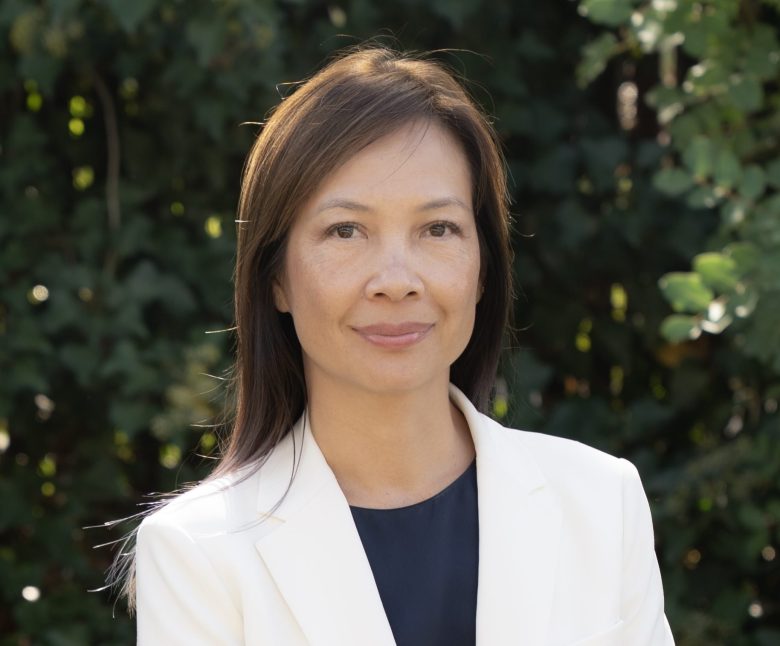 A woman with straight brown hair, wearing a white blazer and dark top, stands outdoors in front of green leafy foliage, looking at the camera with a neutral expression.