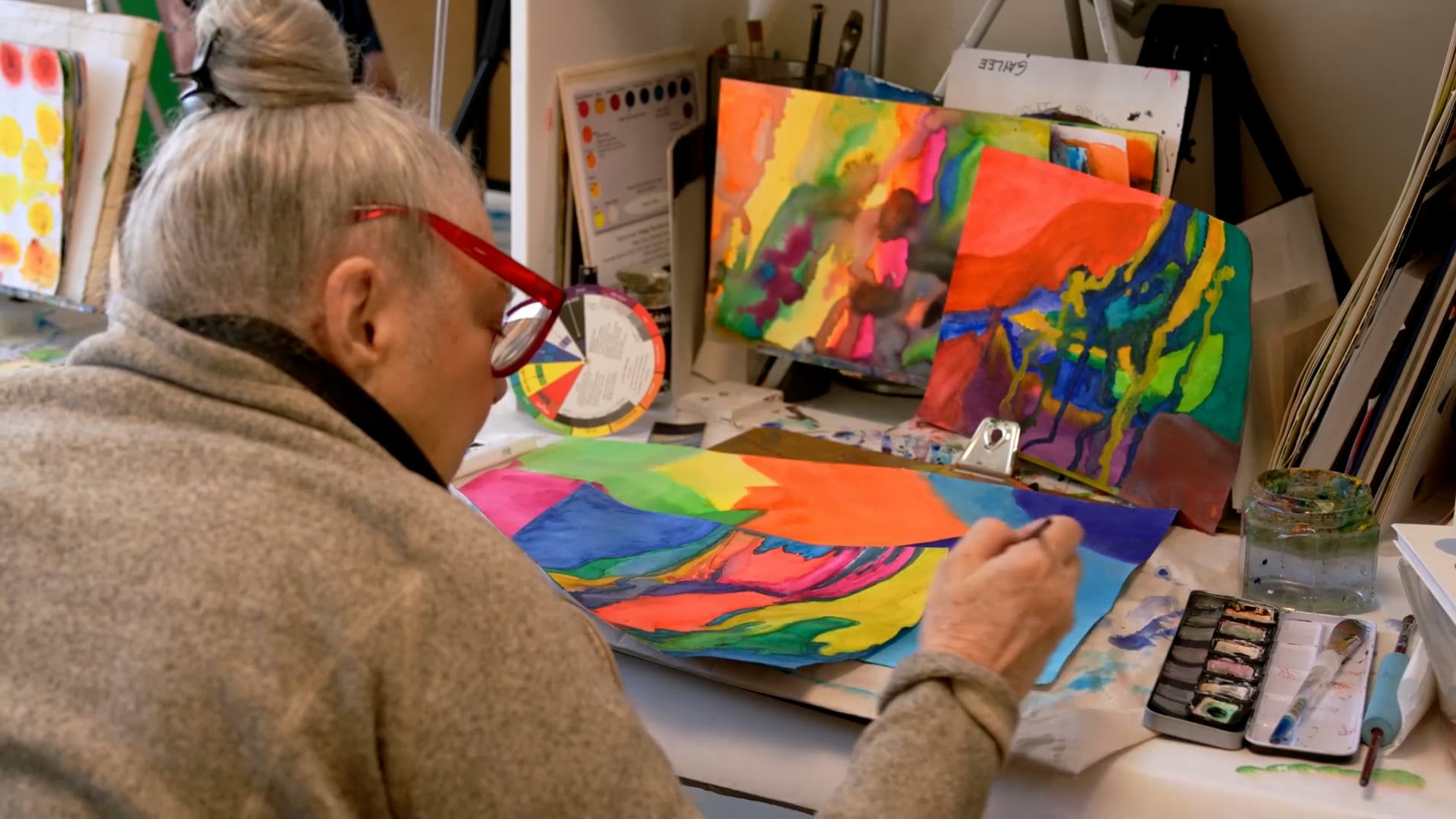 An older woman with gray hair in a bun and red glasses paints a colorful abstract artwork at a desk, with more vibrant abstract paintings and art supplies visible around her.